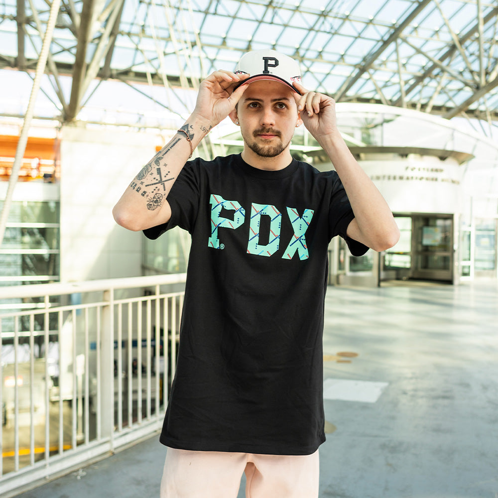 A person with tattoos on their right arm is standing indoors, adjusting their white cap with a black "P" logo. They are wearing an Essential PDX Carpet Tee in black, featuring the limited edition design in large, turquoise letters. The background showcases a glass ceiling and metal railings.